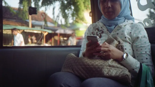 Une femme portant un foulard consulte son smartphone, assise près d'une fenêtre