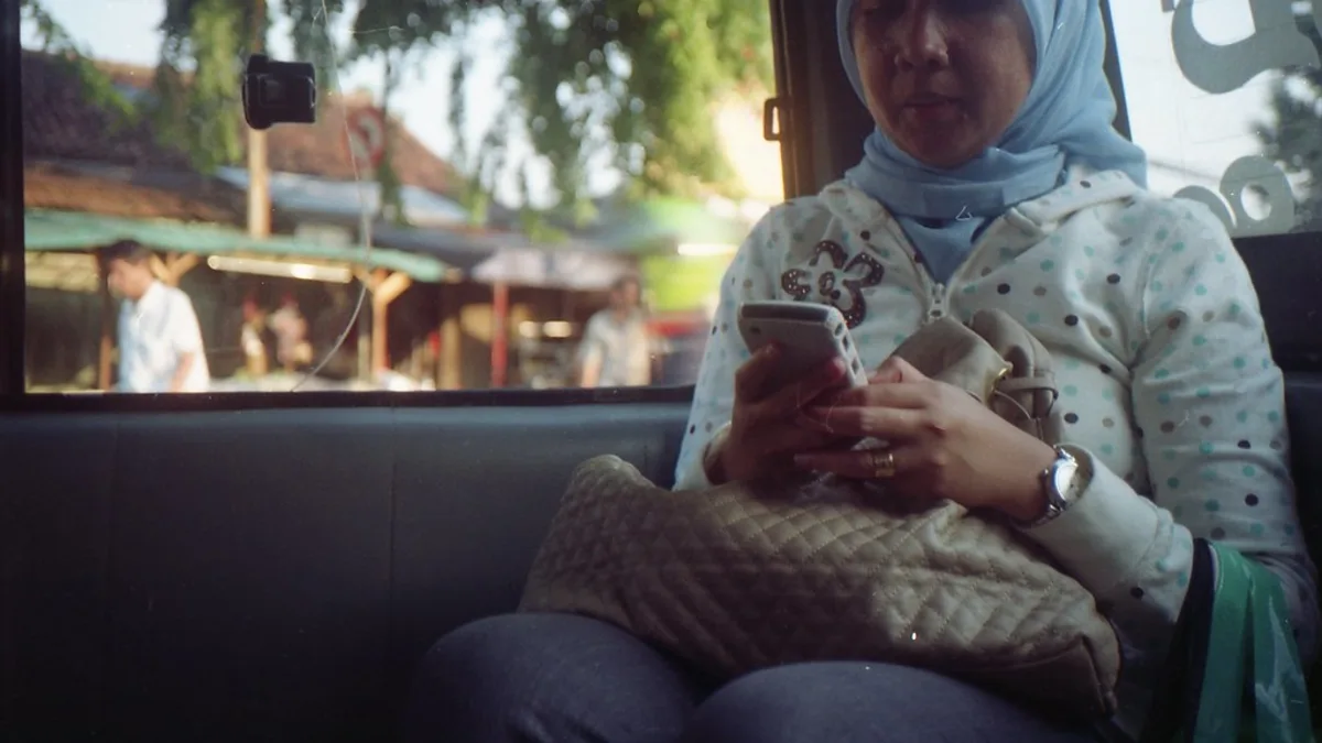 Une femme portant un foulard consulte son smartphone, assise près d'une fenêtre