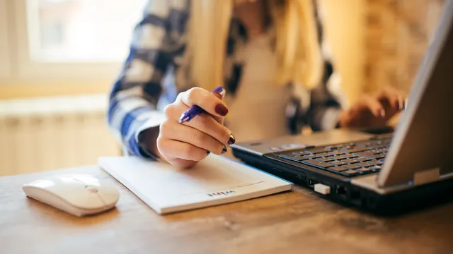 Une femme prend des notes sur un carnet à côté de son ordinateur portable, sur un bureau en bois
